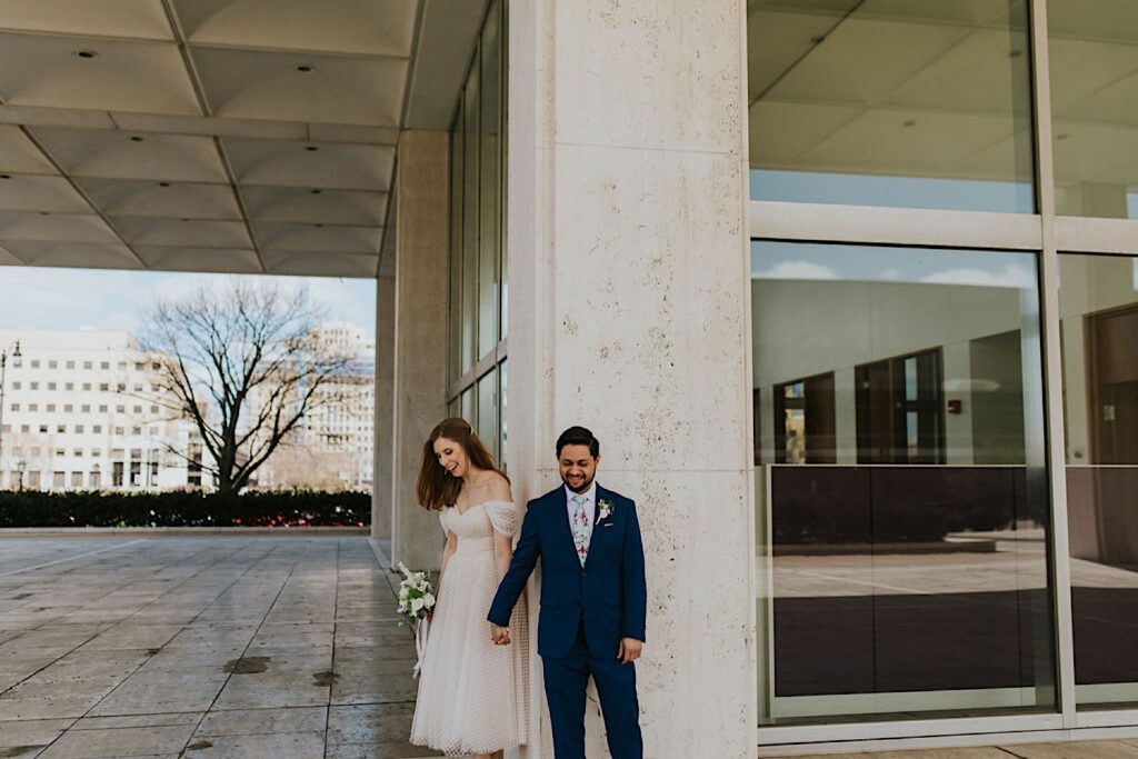 A bride and groom stand next to one another and hold hands around a corner outside of the Horace Mann building in Springfield Illinois