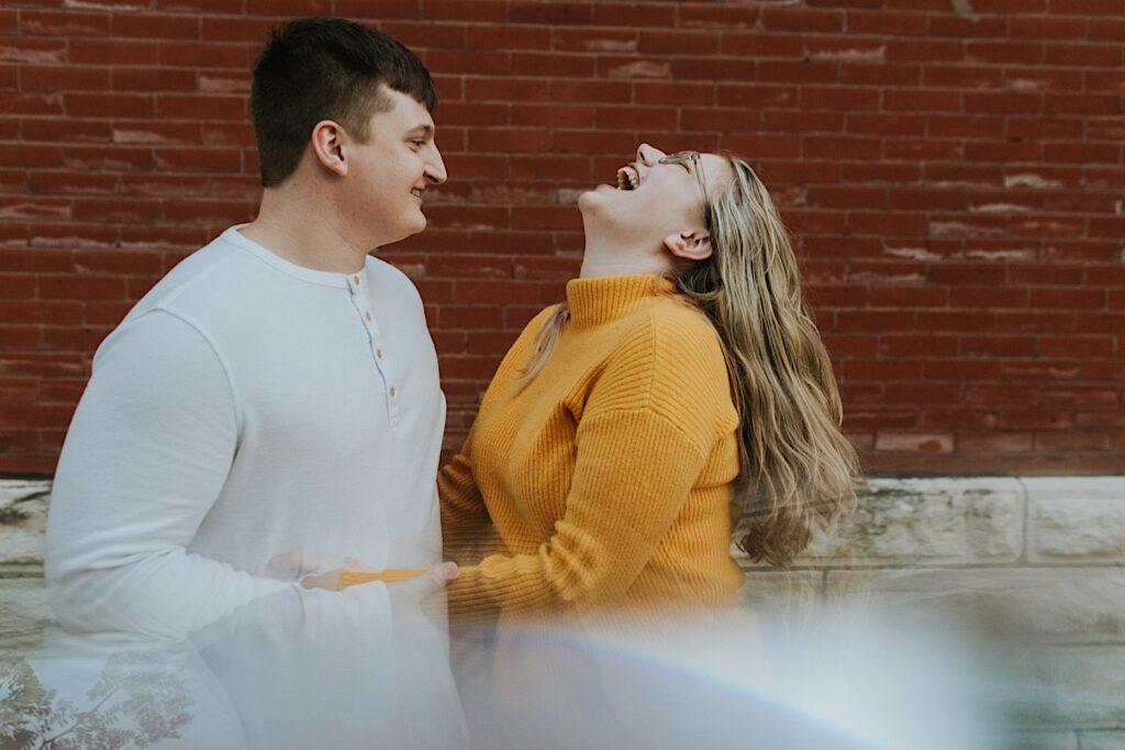 During an engagement session in downtown Springfield, Illinois a woman laughs at the sky as a man smiles at her and holds her hands while they stand together in front of a brick wall