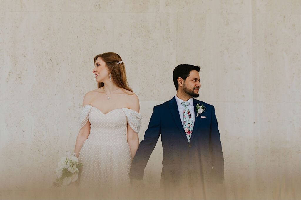 A bride and groom stand in front of a wall and hold hands while facing the camera and looking in opposite directions of one another