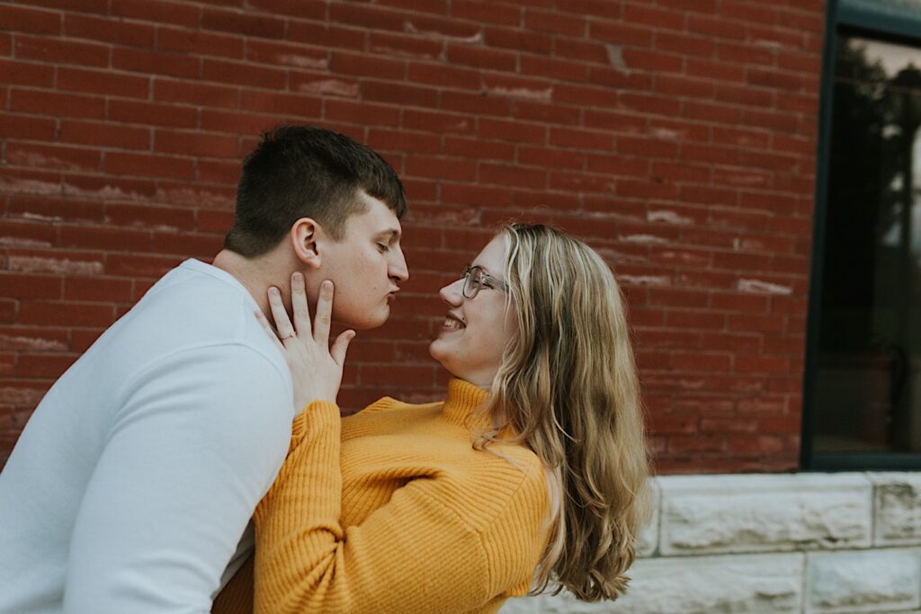 During their engagement session in downtown Springfield, Illinois a man leans in for a kiss as the woman smiles at him, the background is a brick wall