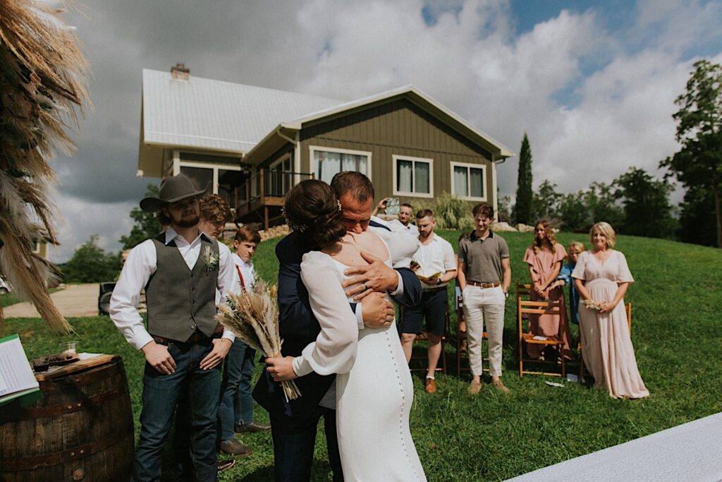 A bride hugs her father after walking down the aisle during her intimate destination wedding in Tennessee, the groom stands next to them and watches while guests stand in the background