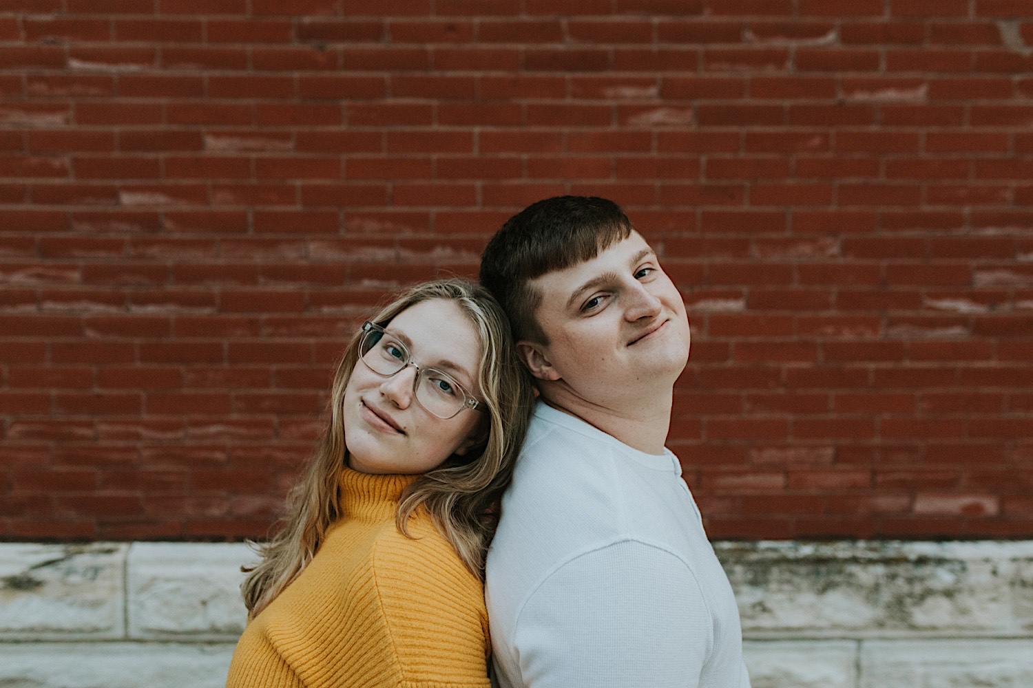 A man and woman stand back to back and smile at the camera while standing in front of a brick wall during their engagement session in downtown Springfield, Illinois