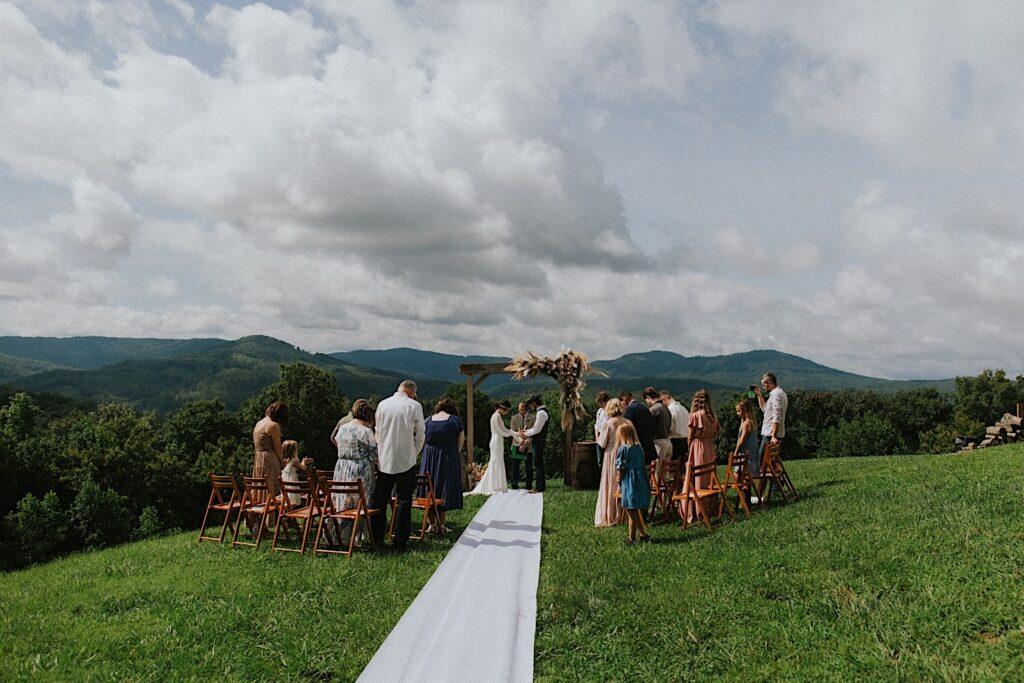 A bride and groom stand holding hands during their intimate destination wedding ceremony in Tennessee, guests stand and watch and in the background are mountains in the distance