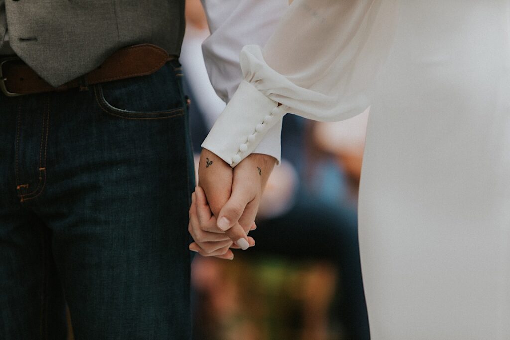 Close up photo of a bride and groom's hands holding one another, each hand has a letter of the other person's initials on them