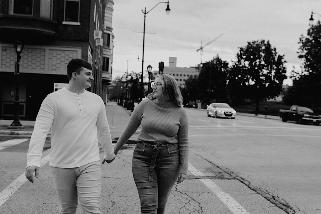 Black and white photo of a couple holding hands and looking at one another while walking across a street during their engagement session in downtown Springfield Illinois