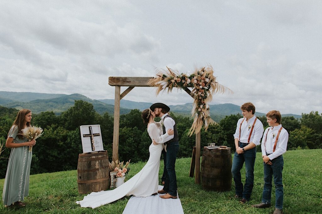 A bride and groom kiss after the ceremony of their intimate destination wedding in Tennessee, behind them are mountains in the distance and their wedding parties watch on either side of them