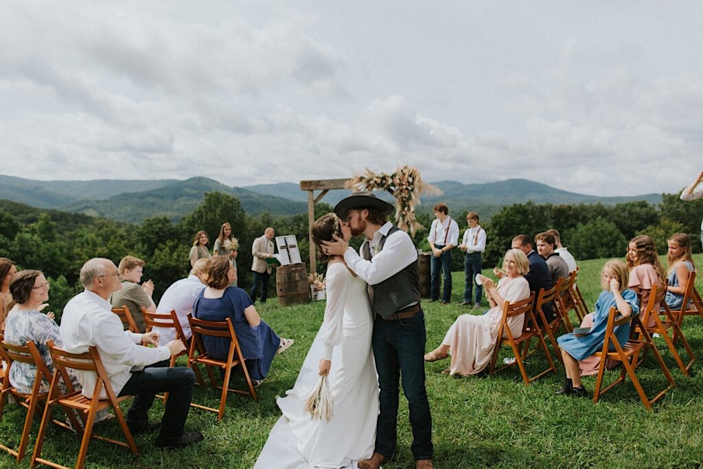 A bride and groom kiss in the middle of the aisle with their guests around them as they exit the ceremony of their intimate destination wedding in Tennessee, behind them are mountains in the distance