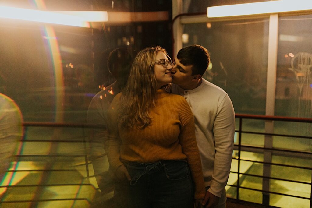 During their engagement session in downtown Springfield, Illinois a woman smiles as a man kisses her from behind while they stand under a well lit clear overhang