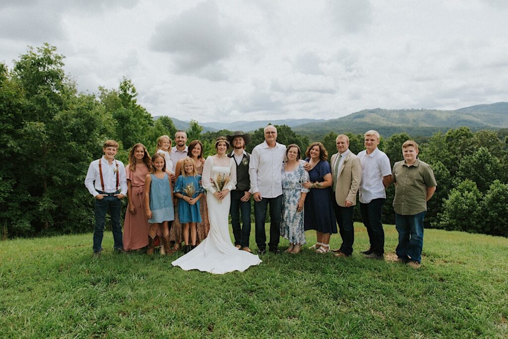 A bride and groom stand with their families on either side of them and smile at the camera after their intimate destination wedding in Tennessee, behind them are mountains in the distance