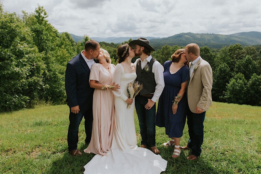 A bride and groom kiss one another, on either side of them are their parents who also kiss one another after their intimate destination wedding in Tennessee, behind the group are mountains in the distance