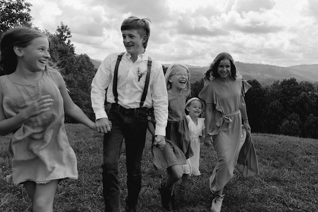 Black and white photo of children all holding hands and running to the camera while laughing, they're guests of an intimate destination wedding in Tennessee and behind them in the distance are mountains