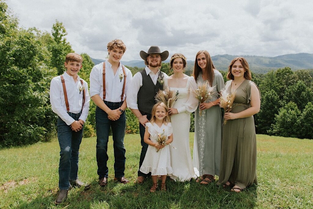 A bride and groom stand with their wedding parties and their flower girl as they all smile at the camera after their intimate destination wedding in Tennessee, behind them are mountains in the distance