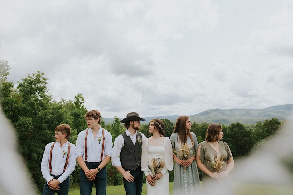 A bride and groom look at one another , on either side of them are their wedding parties who look in opposite directions, behind the group are mountains in the distance