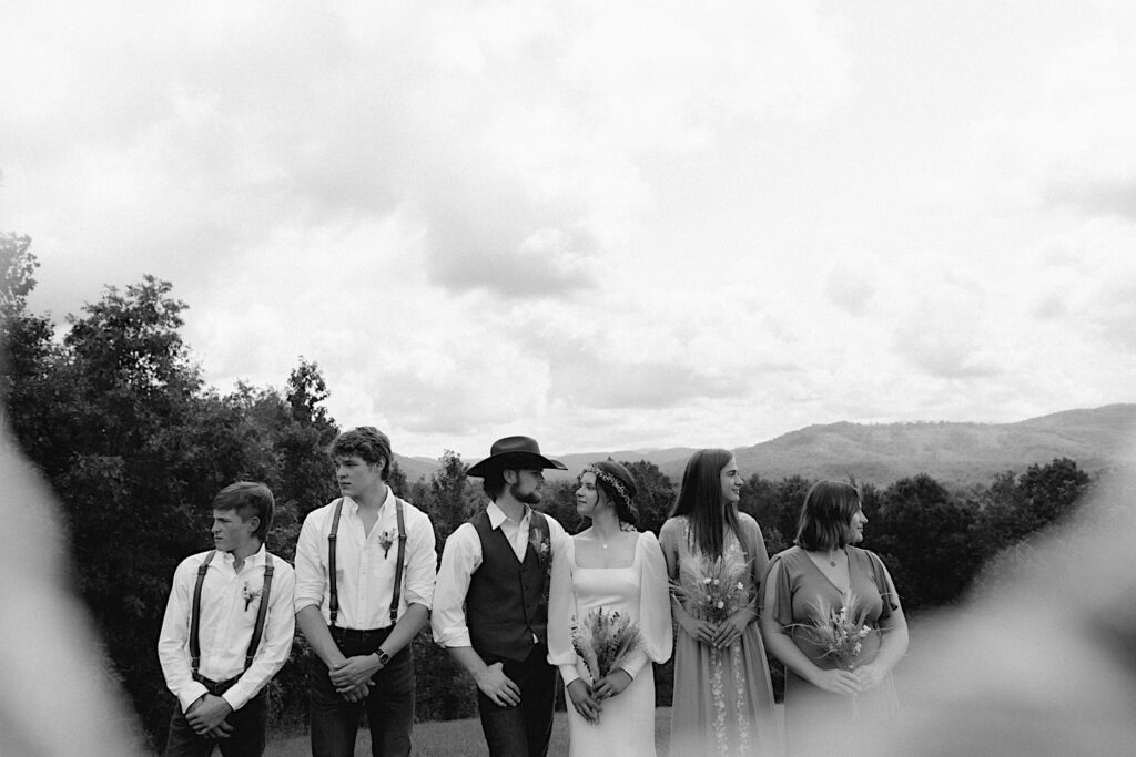 Black and white photo of a bride and groom look at one another , on either side of them are their wedding parties who look in opposite directions, behind the group are mountains in the distance