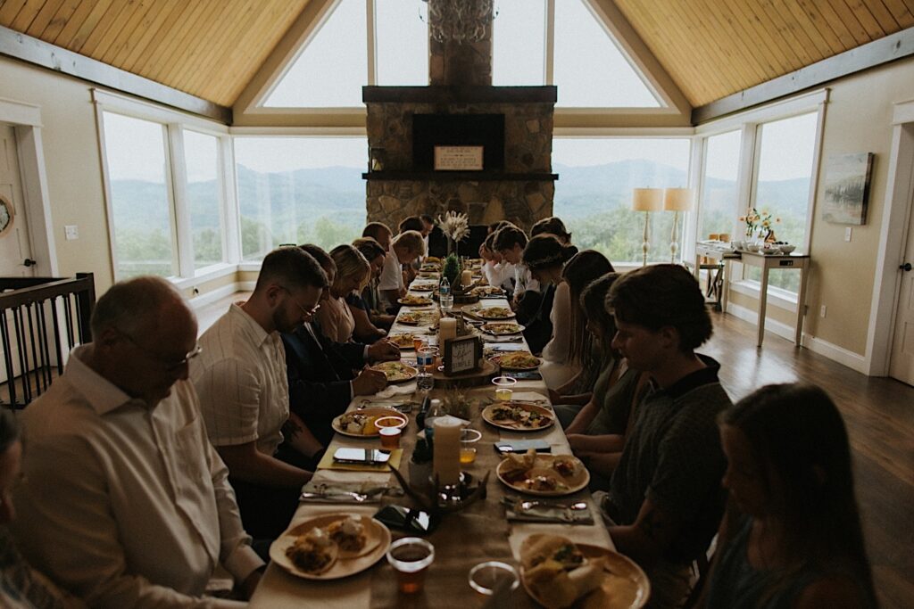 A bride, groom, and guests of their intimate destination wedding in Tennessee all sit at a table indoors and pray before eating their food