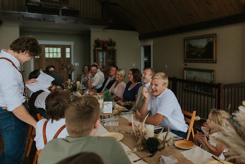 Guests of a wedding laugh while seated at a table as a groomsmen reads a speech while standing
