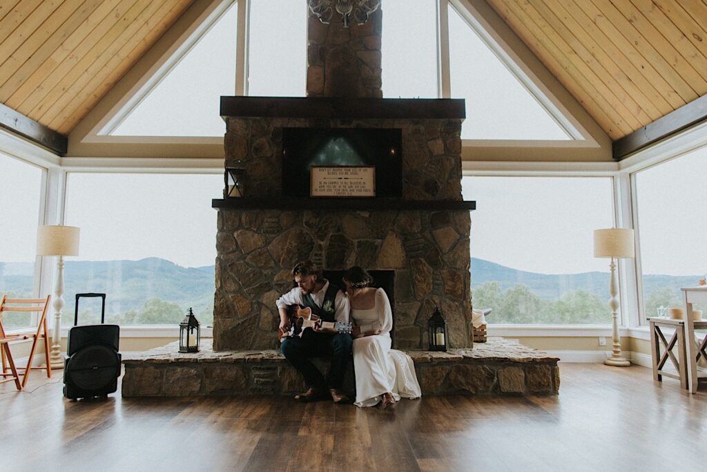 A groom plays an acoustic guitar while sitting at a fireplace with the bride sitting next to him while in an Airbnb during their intimate destination wedding in Tennessee