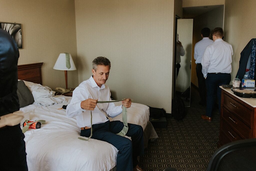 A man sits on a bed looking at a tie as other men in dress shirts exit the hotel room they're in