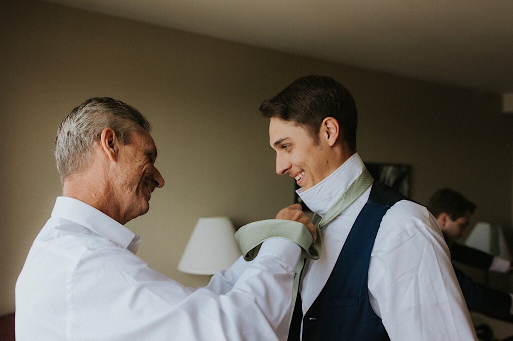 A young groom smiles as his father helps tie his tie on his neck while they get ready for the wedding day