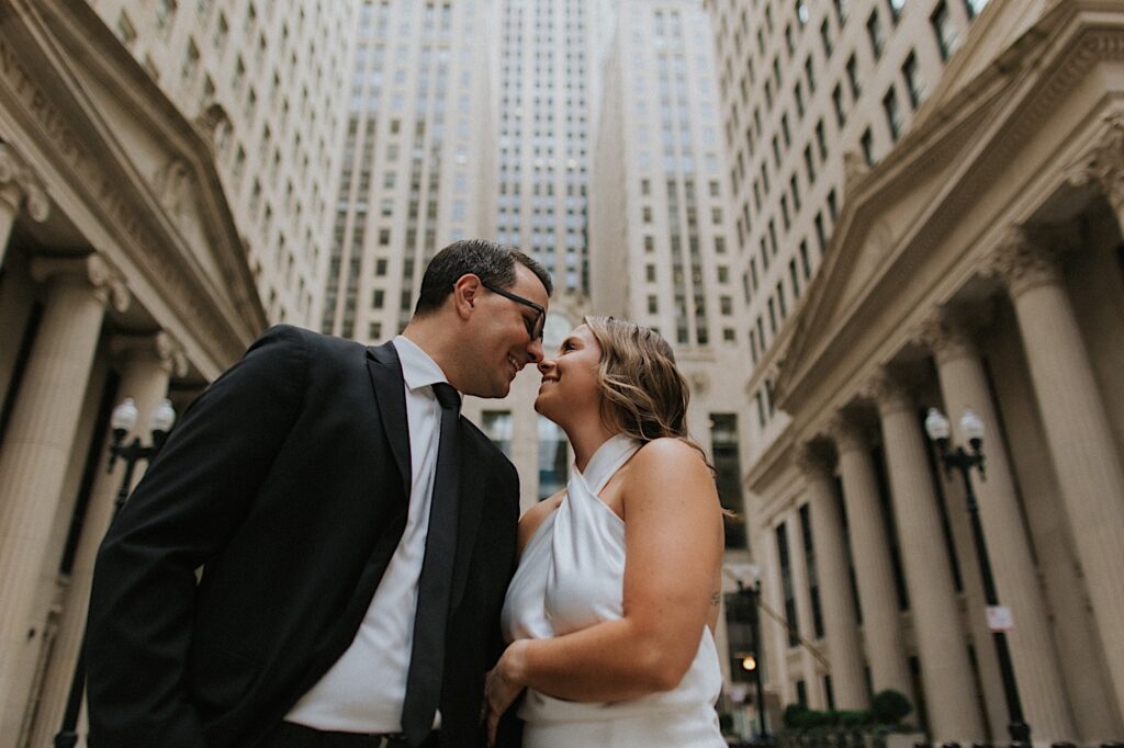 A man and woman smile at one another before kissing while standing in a street of Chicago surrounded by buildings as they take their engagement photos