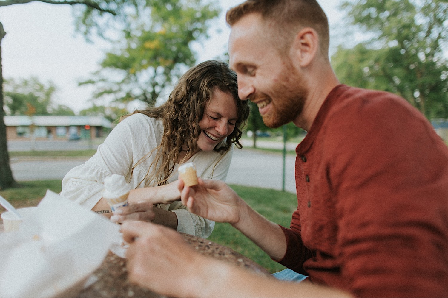 A couple sitting at a table in a park laugh while eating ice cream together during their engagement session in Bloomington-Normal