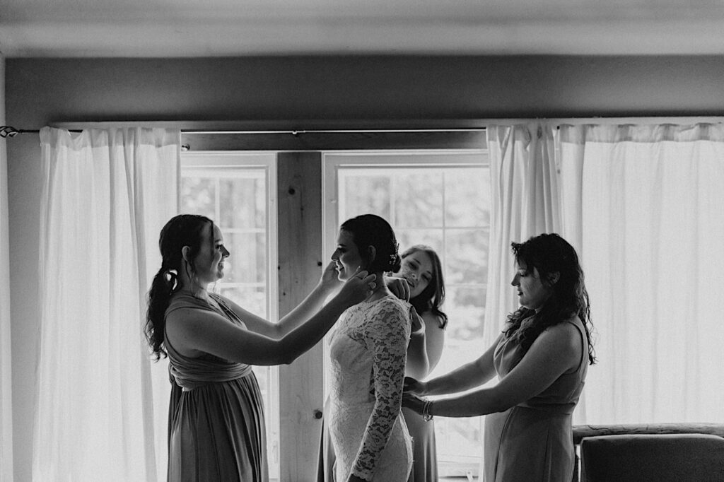 Black and white photo of a bride smiling as her 3 bridesmaids adjust her wedding dress while she stands in front of a window