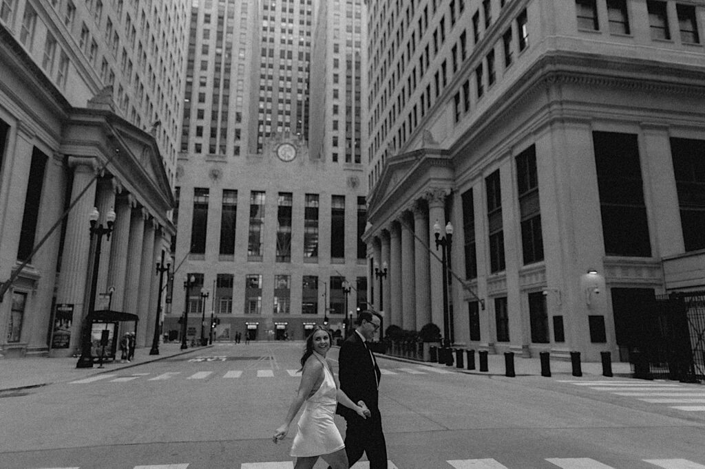 Black and white engagement photo of a couple walking across a street of Chicago together and smiling while holding hands