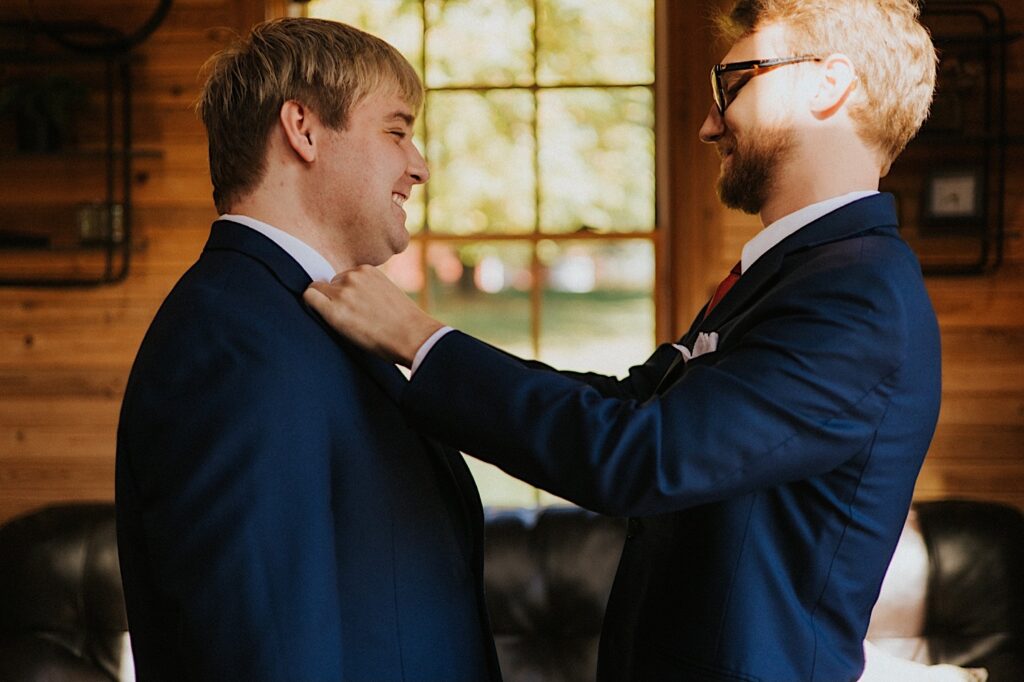 A groom smiles while helping one of his groomsmen adjust his tie as they stand in a wooden room