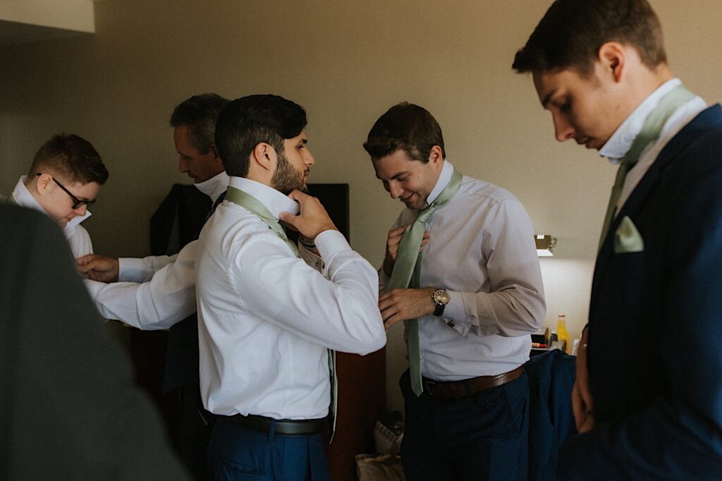 Groomsmen in a hotel room get ready for a wedding day together helping one another put their ties on