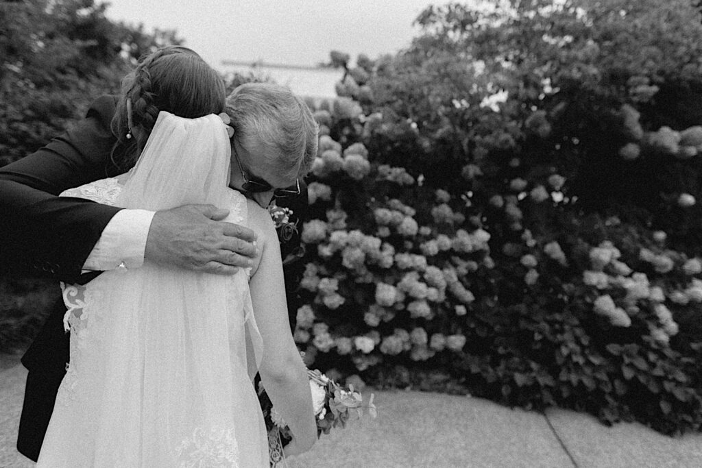 Black and white photo of a father hugging his daughter in her wedding dress, behind them is a wall of flowers