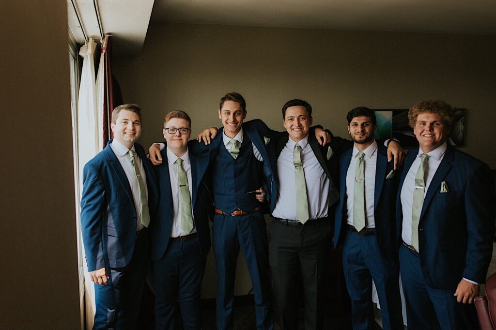 A groom smiles with his 5 groomsmen while standing in front of a window in a hotel room
