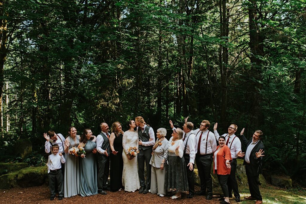 A bride and groom kiss while family members cheer on either side of them while they all stand in front of a forest