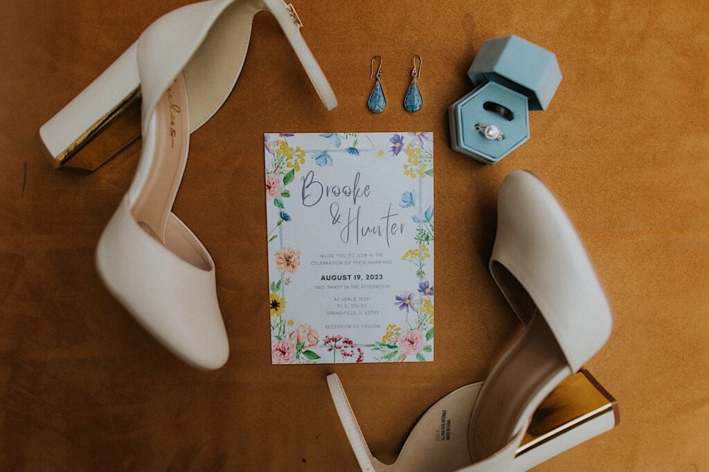 A wedding day flatlay consisting of earrings, wedding rings, women's shoes, and a wedding invite