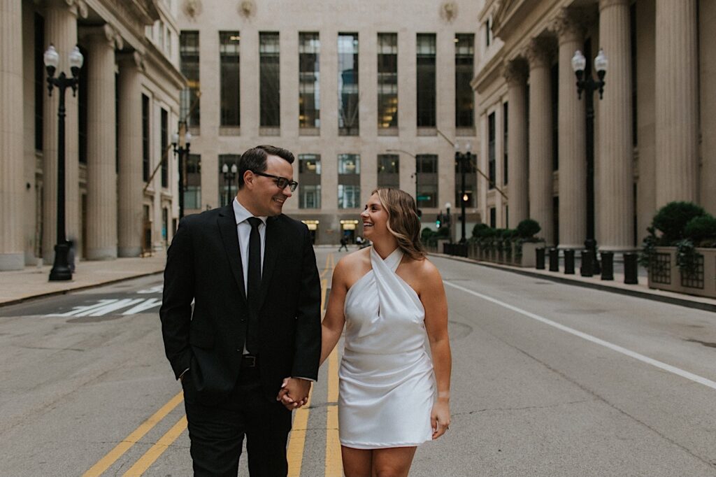 A man and woman walk hand in hand towards the camera and smile at one another while taking engagement photos, they're walking towards the camera down the middle of a street in Chicago