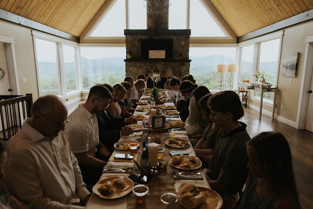 The newlyweds and their guests sit indoors at a long table and pray prior to eating their food 