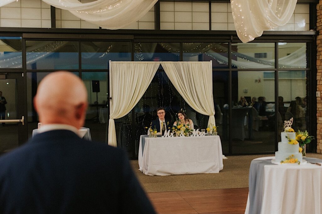 A bride and groom sit at their table during their wedding reception, in the foreground is a man with his back to the camera giving a speech to them. The photo was taken by a Springfield wedding photographer