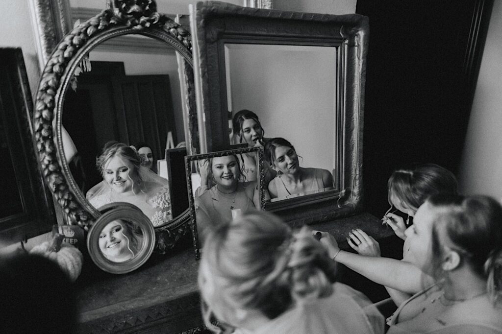 Black and white photo of a bride and her bridesmaids sitting in front of a desk with 4 mirrors on it as they all smile