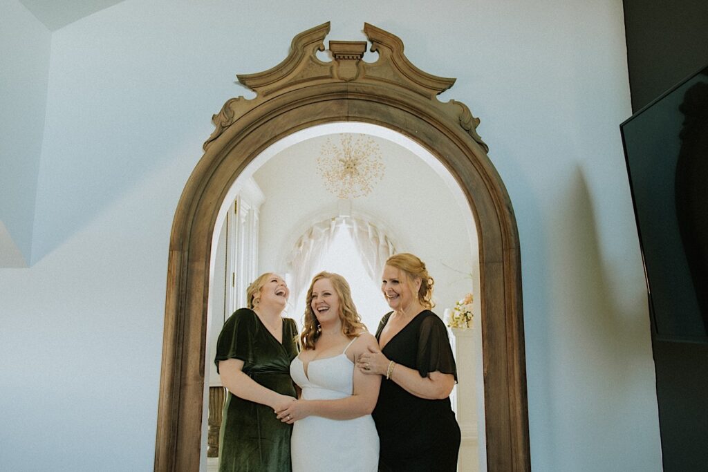 A bride stands with her mother and a bridesmaid in front of a large mirror as they all laugh together. The photo was taken by a Springfield wedding photographer