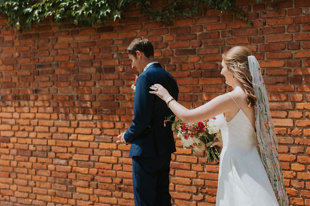 A bride reaches out to tap the shoulder of the groom who is facing away before their first look, behind them is a brick wall