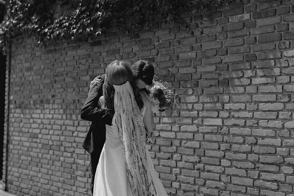 Black and white photo of a bride and groom hugging in front of a brick wall