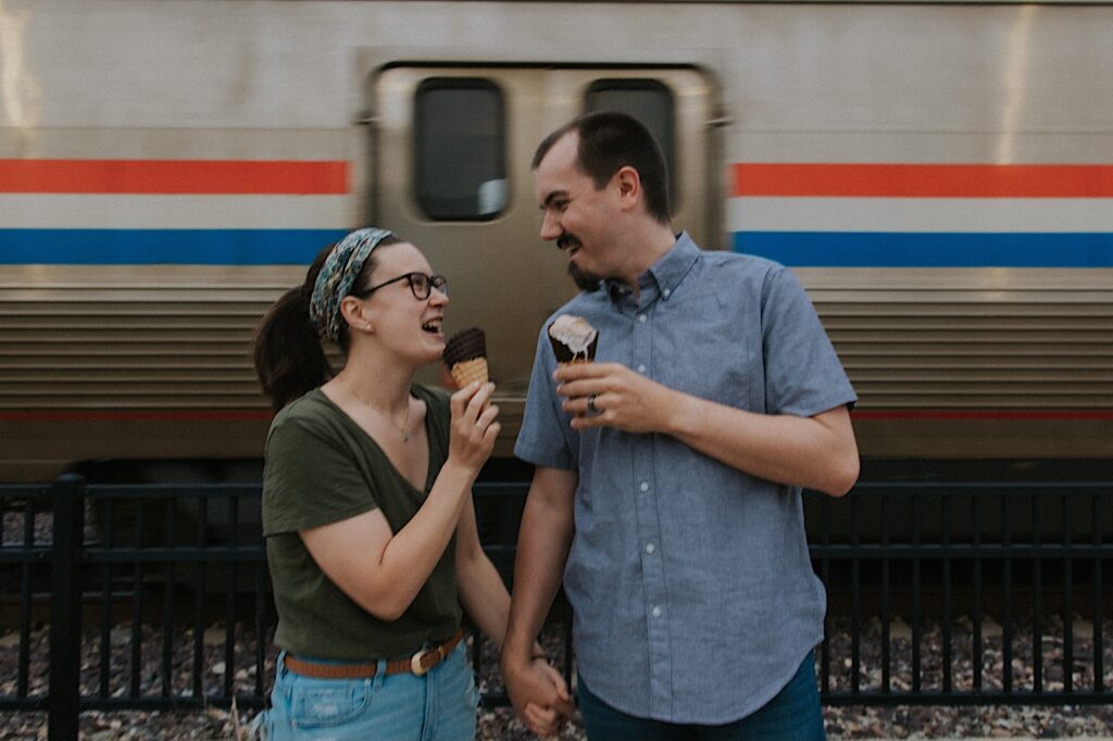 An engaged couple stand holding ice cream and smile at one another while a train passes behind them