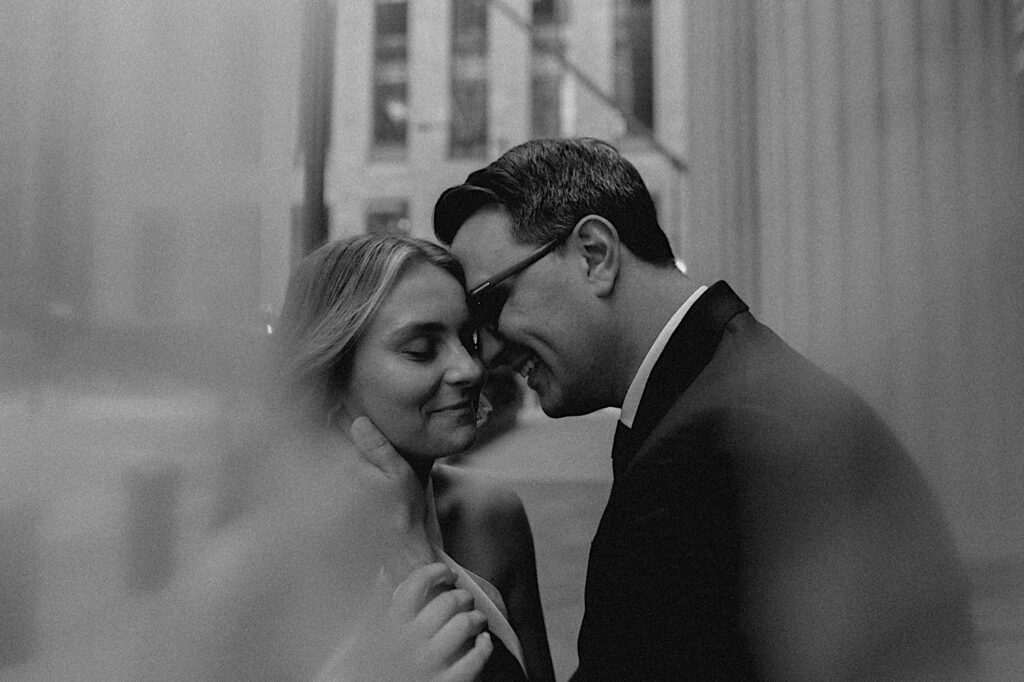Black and white photo of a couple smiling while touching foreheads together as they stand on a street of Chicago while taking engagement photos