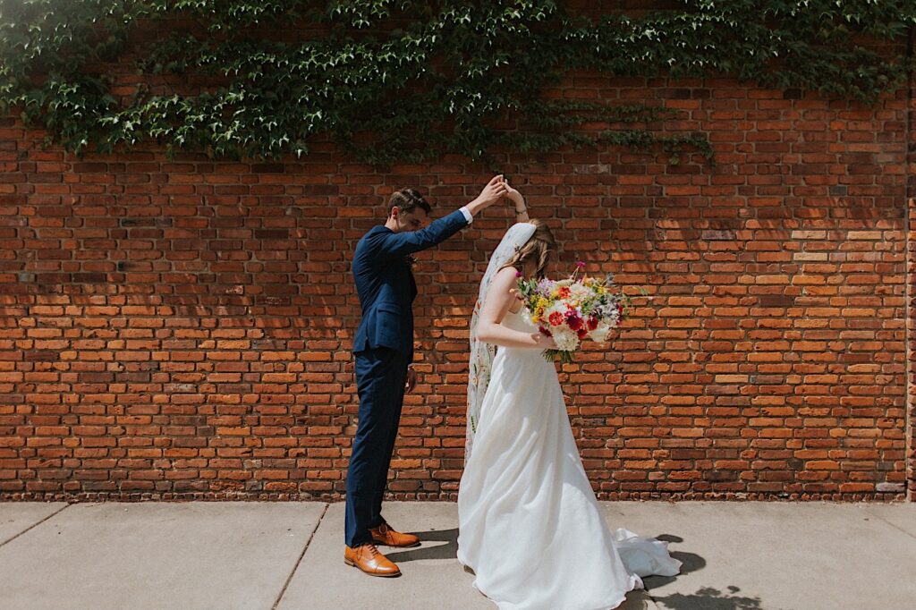 A bride and groom dance together in their wedding attire while on a sidewalk in front of a brick wall