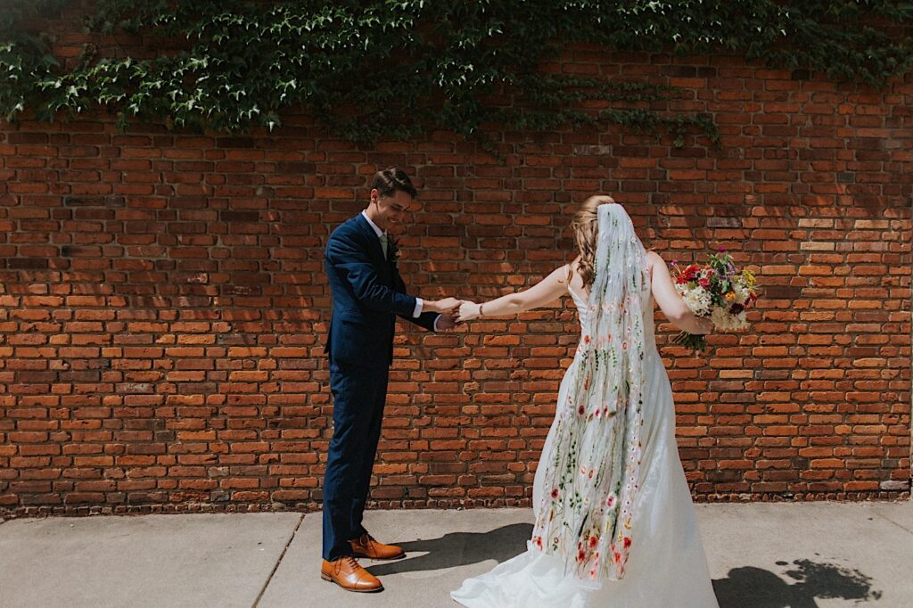 A groom smiles while holding the hand of a bride as she stands in front of him while the two stand in front of a brick wall on a sidewalk together