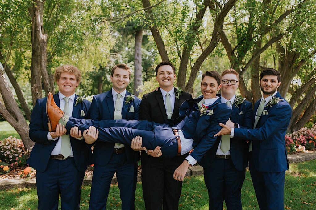 A groom smiles while being held by his 5 groomsmen while they stand outdoors in front of some trees