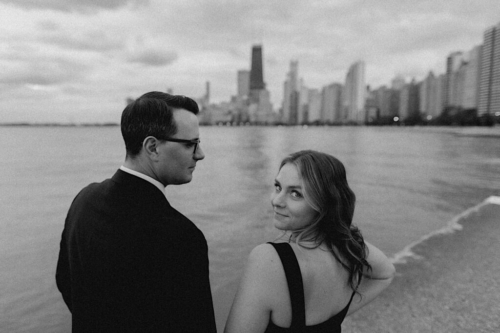 Black and white engagement photo of a couple standing at North Avenue Beach with the Chicago skyline in the background, the man is looking at the woman while she looks back at the camera
