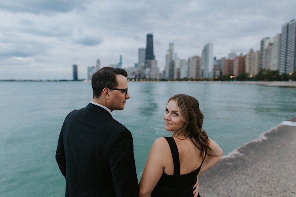 Engagement photo of a couple standing at North Avenue Beach with the Chicago skyline in the background, the man is looking at the woman while she looks back at the camera