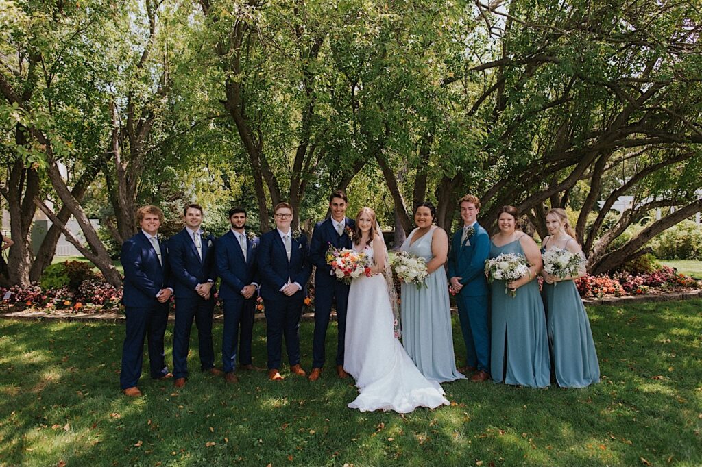 A bride and groom smile while standing with the members of their wedding parties on either side of them while in  front of a row of trees in a park