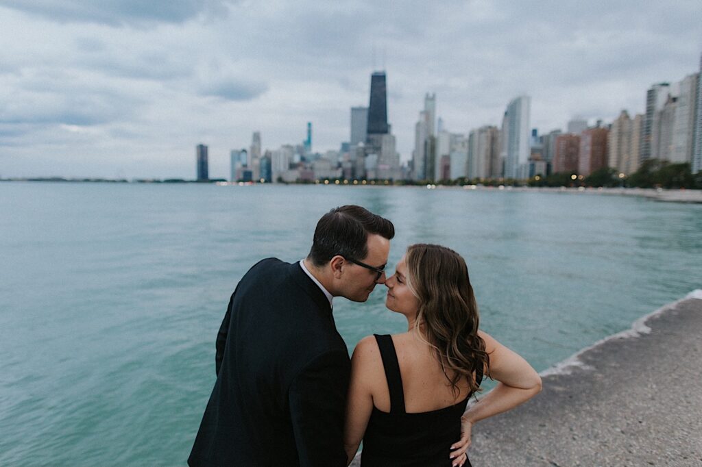 A couple lean in to kiss one another while taking engagement photos at North Avenue Beach, in the background is Lake Michigan and the Chicago skyline
