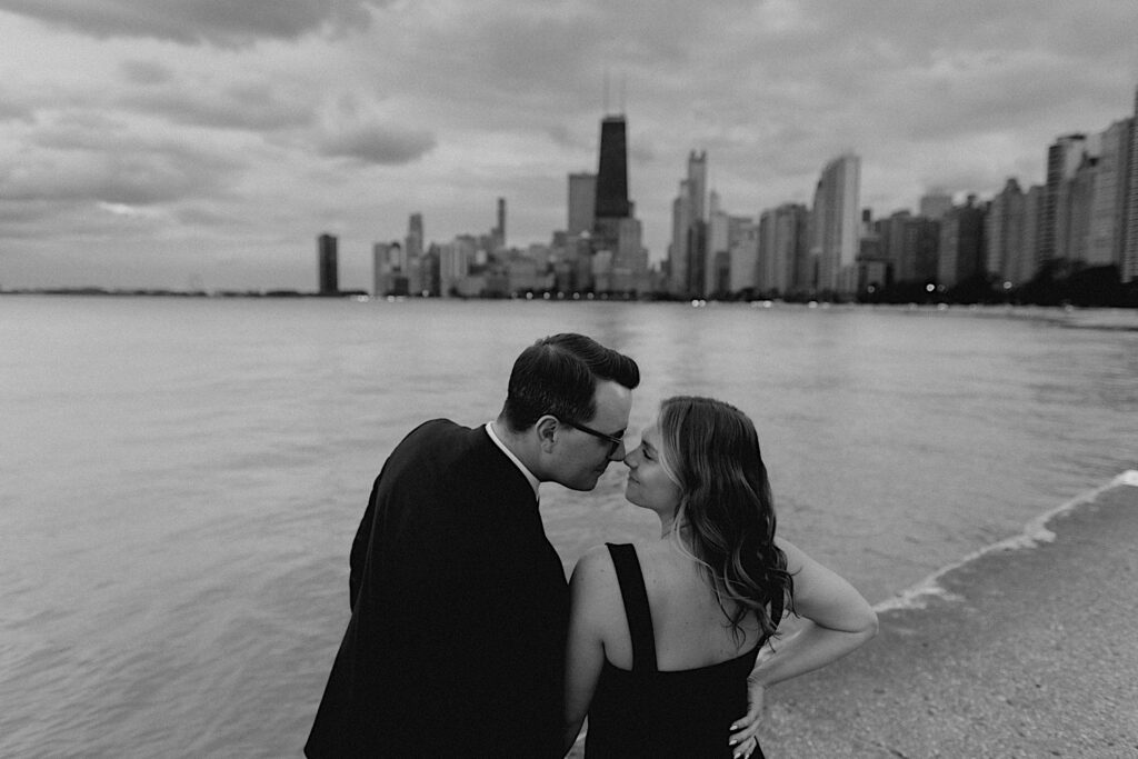 Black and white photo of a couple leaning in to kiss one another while taking engagement photos at North Avenue Beach, in the background is Lake Michigan and the Chicago skyline